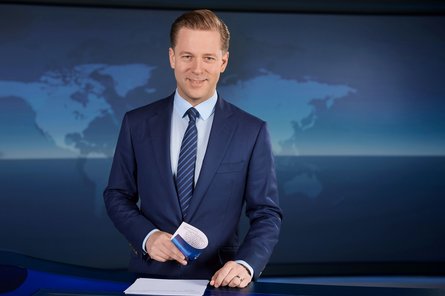 Ein lächelnder Mann in Anzug und Krawatte in einem Nachrichtenstudio