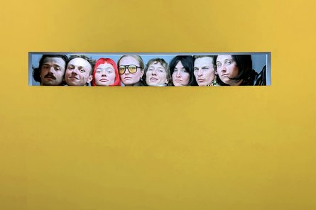 Verletschungen 8 Personen schauen nebeneinander durch ein breites Fenster in einer gelben Wand