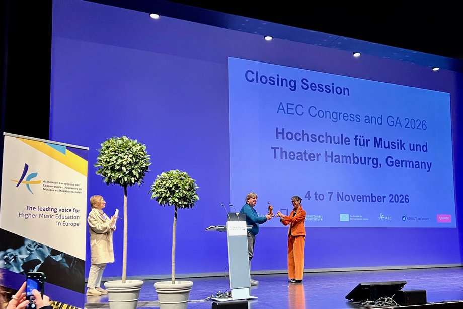 Abschlussveranstaltung AEC Salzburg Drei Personen stehen auf einer Bühne und im Hintergrund ist eine Leinwand